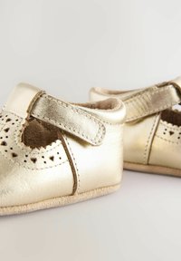 Auksiniai odiniai batukai kūdikiams su perforuotais raštais, reguliuojama sagtimi ir minkštomis velveto pado detalėmis. Apvalus priekis ir tekstūruotas vidinis apdailos paviršius.