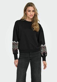 Črna dukserica širokega kroja z dekorativnimi vzorčenimi rokavi v beli, rdeči in zeleni barvi. Ribu ano konci in rob poudarjajo obliko.