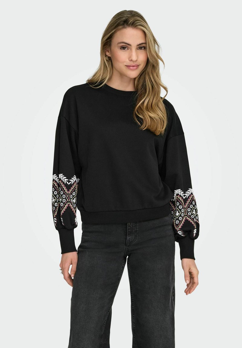 Črna dukserica širokega kroja z dekorativnimi vzorčenimi rokavi v beli, rdeči in zeleni barvi. Ribu ano konci in rob poudarjajo obliko.