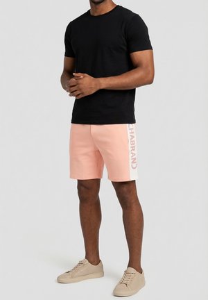 Uomo che indossa una maglietta nera, pantaloncini rosa chiaro con striscia laterale bianca e scritta, e scarpe da ginnastica beige, in piedi con le mani intrecciate.