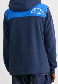 Persona con una giacca in pile blu navy con uno yoke e cappuccio blu brillante, e un logo semi-circolare bianco sulla spalla posteriore.