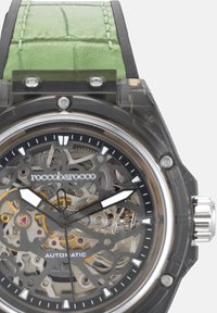 Orologio con cassa trasparente, dettagli argentati, quadrante scheletrato nero, movimento automatico, cinturino in pelle verde con motivo coccodrillo.
