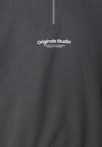 Felpa grigia scura con zip a quarti e testo bianco "originals Studio, portato a te da Jack & Jones, progettato a Copenhagen" sul petto.