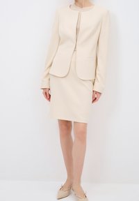 Blazer beige ajustado y falda hasta la rodilla hecha de tejido texturizado. El blazer tiene un escote redondo y dos bolsillos en la parte delantera; acabado suave.