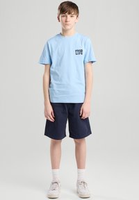 Light blue cotton t-shirt with "GOOD LIFE" printed on the chest, paired with navy shorts. Worn with white socks and sneakers.