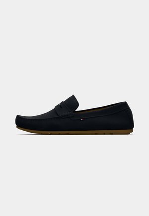 Mocassin en cuir noir avec un bout arrondi, détail de bandeau à pièce, et semelle en caoutchouc marron, présenté en profil sur un fond blanc.