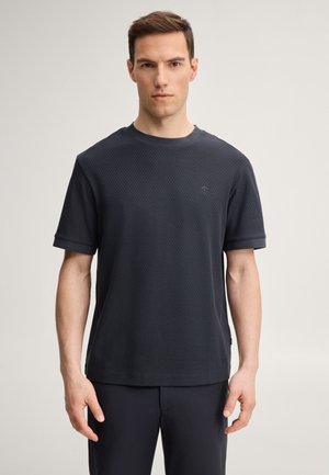 Uomo in piedi contro uno sfondo semplice, indossa una camicia scura a maniche corte con trama e pantaloni coordinati, guarda dritto avanti con un'espressione neutra.