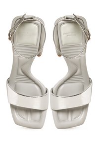 Sandale argintii cu baretă la gleznă, vârf pătrat, talpă căptușită și curea frontală lată și lucioasă, cu numele brandului gravat pe tălpici.