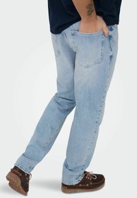 Man draagt lichtblauwe spijkerbroek en donkerbruine schoenen, staat met één hand in zijn achterzak en een tatoeage zichtbaar op de onderarm.