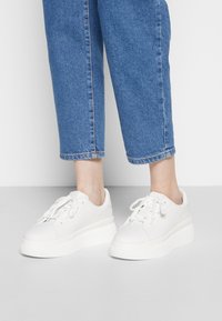 Baskets blanches avec un dessus en cuir lisse, bout arrondi et semelle épaisse texturée. Des lacets et un minimum de détails de couture sont présents.
