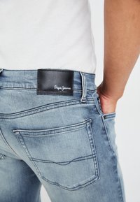Vaqueros de denim azul claro con un parche de cuero negro etiquetado "Pepe Jeans London", que cuenta con bolsillos traseros y un diseño ajustado.