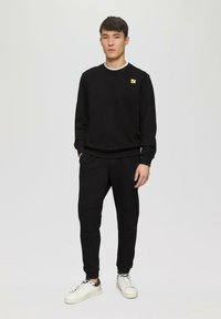 Schwarzer Kapuzenpullover mit gelbem Logo, kombiniert mit schwarzen Jogginghosen mit strukturierten Einsätzen und weißen Sneakers mit grauen Akzenten.