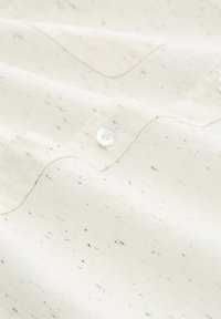 Hellbeiges Hemd mit gesprenkelter Textur, das eine Brusttasche und weiße Knöpfe besitzt. Der Stoff weist ein dezentes, gewebtes Muster auf.