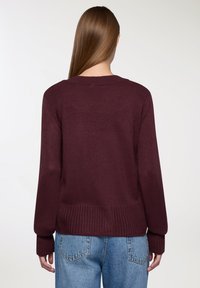Burgundowy sweter z dzianiny z długimi rękawami i ściągaczami. Posiada szeroki dekolt i ściągacz u dołu. Noszony z jasnoniebieskimi dżinsami.