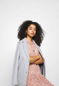 Lichtgrijze wollen jas over een roze bloemenjurk, met een keyhole-halslijn en korte mouwen. Het model heeft krullend haar en opvallende oorbellen.