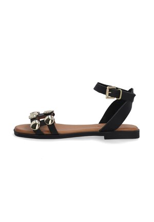 Black flat sandal with adjustable ankle strap, gold circular accents, and a brown sole. Smooth textile straps create a minimalist design.