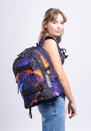 Junge Frau in ärmellosem Top und Jeans mit großem lila-orange gemustertem Rucksack, seitlich stehend vor weißem Hintergrund.