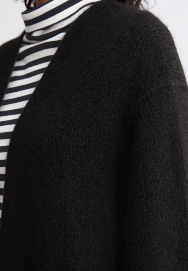 Schwarze, strukturierte Strickjacke aus weichem Garn, getragen über einem schwarz-weißen gestreiften Rollkragenpullover. Die Strickjacke hat eine lockere Passform und eine offene Vorderseite.