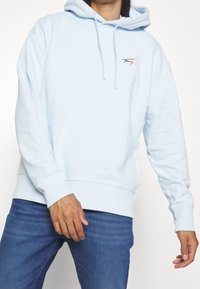 Sudadera con capucha azul claro hecha de mezcla de algodón, con un logo frontal, capucha con cordón y puños y dobladillo acanalados. Se combina con vaqueros azul oscuro.