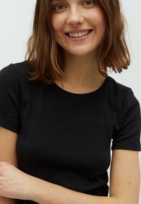Jauna sieviete ar plecu garuma brūnajiem matiem, tērpusies melnā īsā piedurknē, smaida pret vienkāršu gaišu fona.