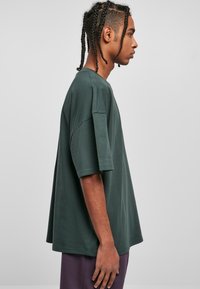 Tricou oversized de culoare verde închis, confecționat dintr-un material moale, cu mâneci scurte și decolteu rotund, prezentat din lateral pe un fond neutru.