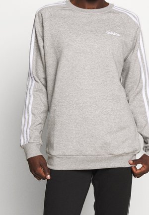 Person trägt einen grauen Adidas-Pullover mit weißen Streifen an den Ärmeln und schwarze Hosen, die Hände passen den Saum des Pullovers an.