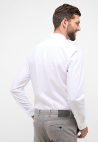 Camicia bianca a maniche lunghe con colletto classico, texture liscia e polsini con bottoni, indossata con pantaloni grigi dotati di tasche posteriori e dettagli in pelle.