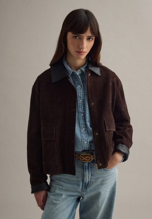 Frau trägt eine dunkelbraune Wildlederjacke über einem hellblauen Jeanshemd, das in eine hellblaue Jeans mit gemustertem Gürtel gesteckt ist, und steht vor einem einfarbigen Hintergrund.