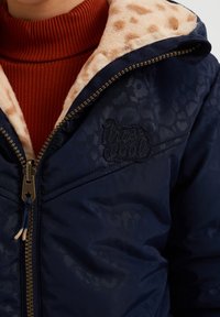 Marineblaue Steppjacke mit einem leichten Leopardenmuster-Futter, ausgestattet mit einem Reißverschluss und einem gestickten Logo auf der Brust. Begleitet von einem roten Rollkragenpullover.
