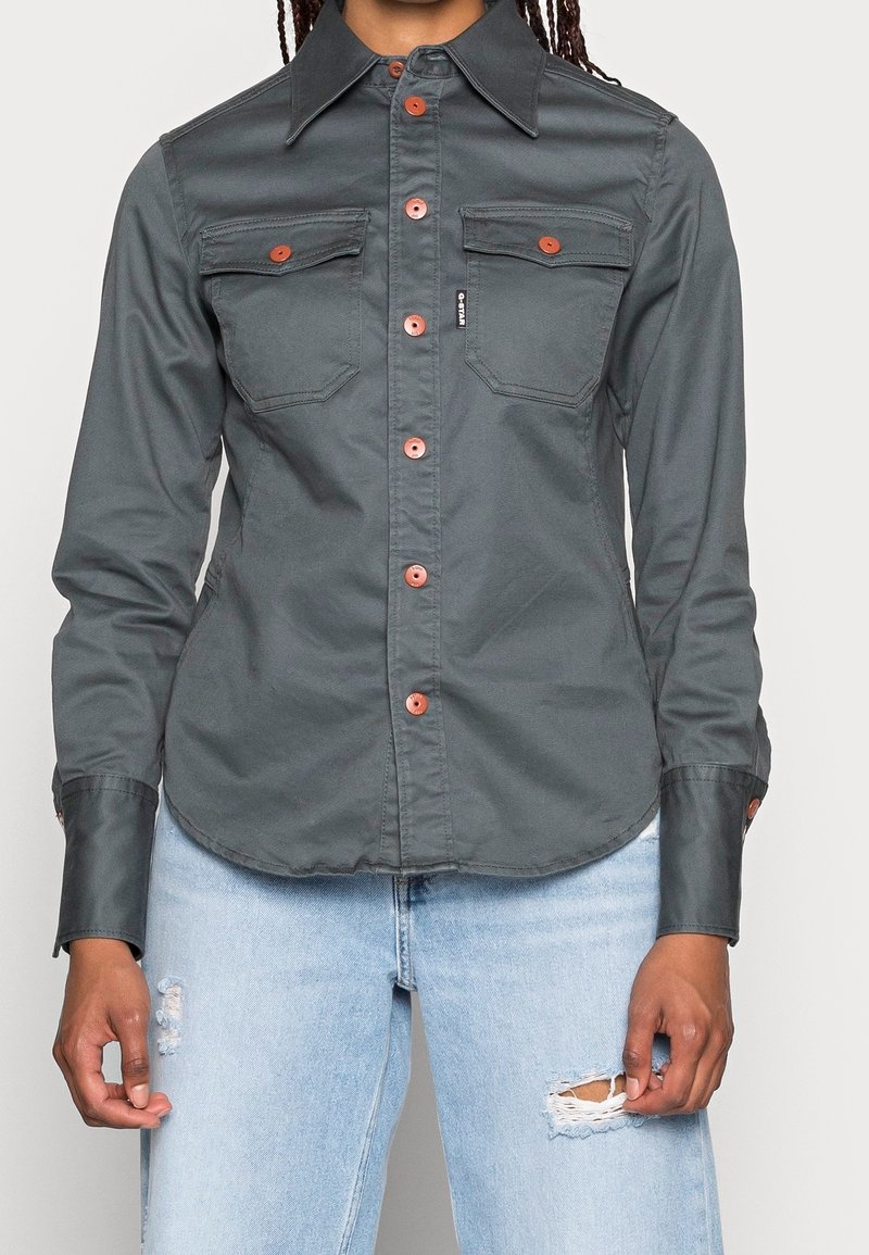 Femme portant une chemise boutonnée gris foncé avec des poches poitrine et un jean bleu clair déchiré, debout devant un fond uni.