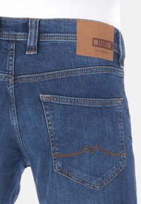 Jeans in denim blu con un'etichetta in pelle marrone sulla tasca posteriore, caratterizzati da cuciture a contrasto e un design curvo della tasca.