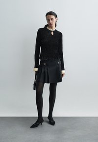 Γυναίκα που φοράει μια μαύρη υφασμάτινη μπλούζα με μακριά μανίκια και άσπρο γιακά και μανσέτες, μαύρη πλισέ φούστα, μαύρες καλσόν, γόβες και κρατάει μια μικρή μαύρη τσάντα.