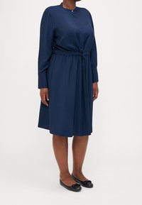 Dunkelblaues langärmliges Kleid mit rundem Halsausschnitt, elastischem Bund und Bindeband. Glattes Material, knielang, kombiniert mit schwarzen Ballerinas.