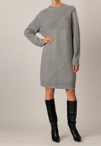 Robe en tricot grise avec un col côtelé et un design asymétrique. Manches longues et ourlet se terminant au milieu de la cuisse. Portée avec des bottes noires montantes.