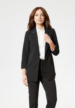 Vrouw met bruin haar, gekleed in een zwarte blazer, witte blouse met knopen en een zwarte broek, staand tegen een eenvoudige lichte achtergrond.