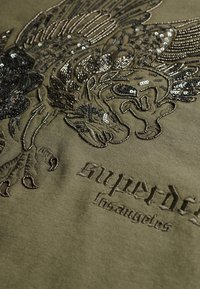 Camiseta verde oliva con un bordado intrincado en negro y lentejuelas metálicas que representan un diseño, con el texto "superdry losangeles".