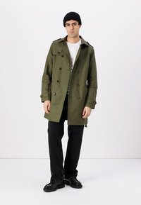 Abrigo color verde oliva con botones dobles, cuello de pico y tela texturizada, combinado con pantalones negros y un gorro.