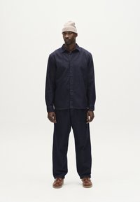 Camisa y pantalones de mezclilla azul marino con un corte relajado, con cierre de botones en la parte delantera. Un gorro de punto color beige y calzado marrón completan el conjunto.
