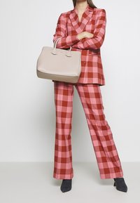 Różowo-czerwony w kratę garnitur z dopasowaną marynarką i szerokimi spodniami, w połączeniu z jasnobeżową, strukturalną torebką i ciemnymi botkami na obcasie.