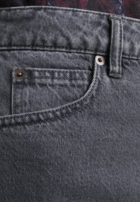 Šedé denimové džíny s přední kapsou, šitými švy a kovovými nýty na kapse, které ukazují texturovaný, opotřebovaný vzhled látky.