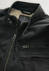 Černá bunda se zipem vpředu, texturovanou podšívkou a několika kapsami na zip. Obsahuje patentky a kontrastní zlatý zip.