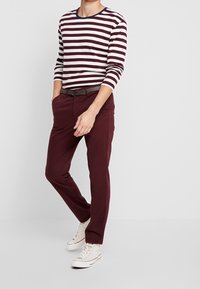 Vinröda byxor med rak bendesign, parat med en vinröd och vit randig långärmad skjorta. Har rund halsringning och ett brunt bälte.