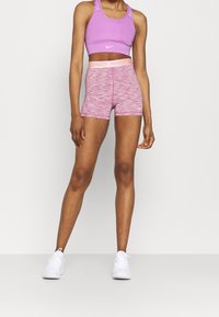 Kobieta w różowym staniku sportowym Nike i dopasowanych, różowych szortach Nike Pro z białymi sneakersami, stojąca na jednolitym tle.
