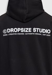 Zwarte hoodie met witte tekst op de achterkant met de tekst "©DROPSIZE STUDIO (030) | CREATIVE VISIONS - AUTHENTIEK ONTWERP | EST 2020".