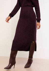 Strikket midilang nederdel i vinrød med kabelmønster, der har høj talje og sideslids, kombineret med brune læderstiletter op til knæet.