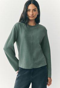 Maglione verde a maniche lunghe con collo a giro, realizzato in tessuto texturizzato, con dettagli delle cuciture lungo il davanti, abbinato a jeans scuri. Design semplice e moderno.