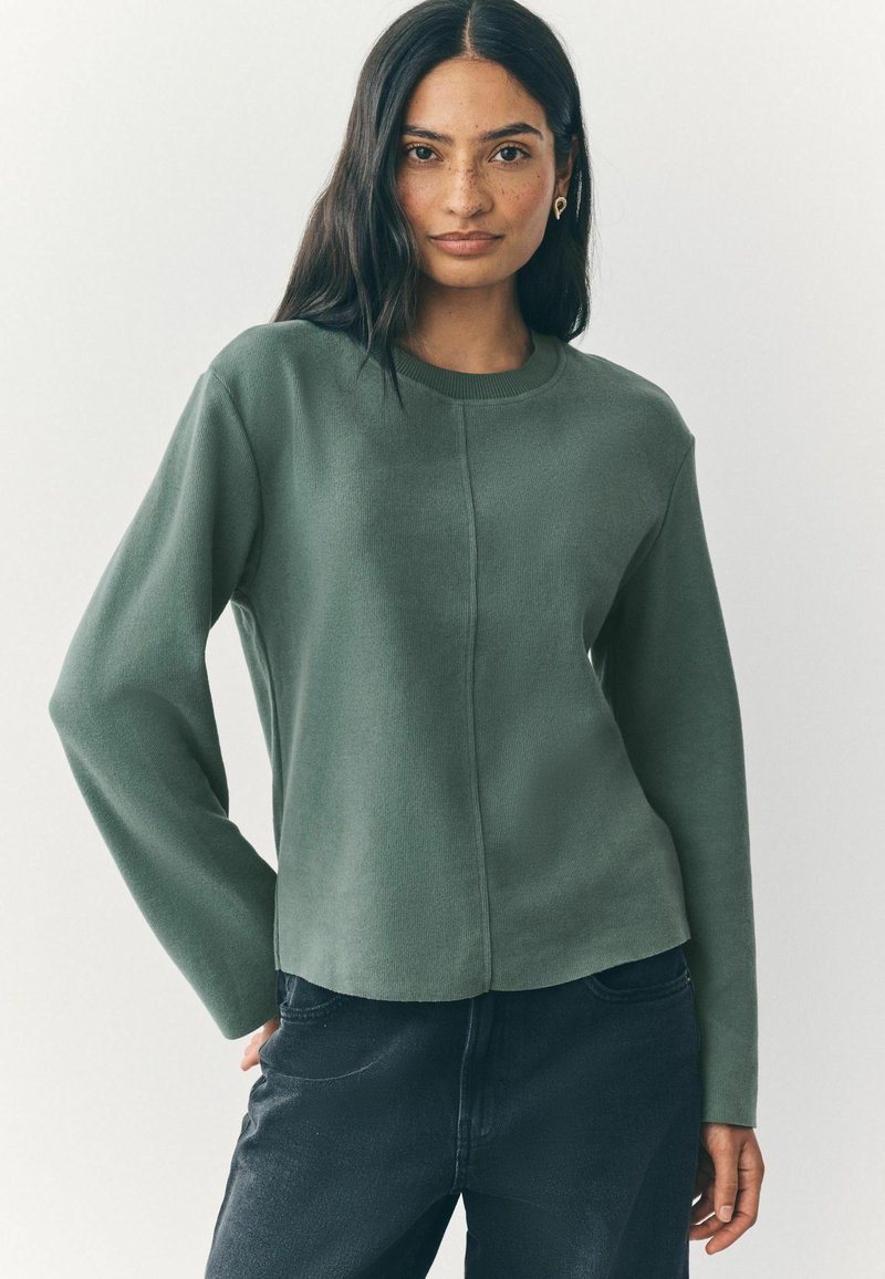 Green long-sleeve crew neck top, textured fabric, seam detailing down the front, paired with dark jeans. Simple and modern design.