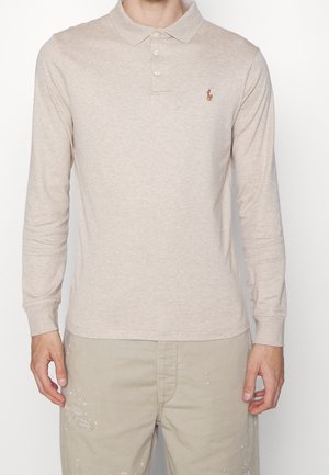 Homme portant un polo à manches longues beige avec trois boutons et un pantalon kaki clair avec des éclaboussures de peinture, debout devant un fond blanc.