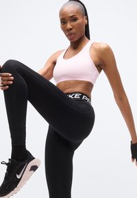 Αθλητικό σουτιέν σε ανοιχτό ροζ, κολάν συμπίεσης σε μαύρο με ζώνη μέσης με μοτίβο. Το μοντέλο δείχνει θέση με ψηλό γόνατο, φορώντας μαύρα αθλητικά παπούτσια.