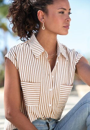 Camisa de manga corta a rayas en algodón crema y marrón claro con dos bolsillos frontales; collares en capas de tono dorado y pendientes de aro complementan el look.